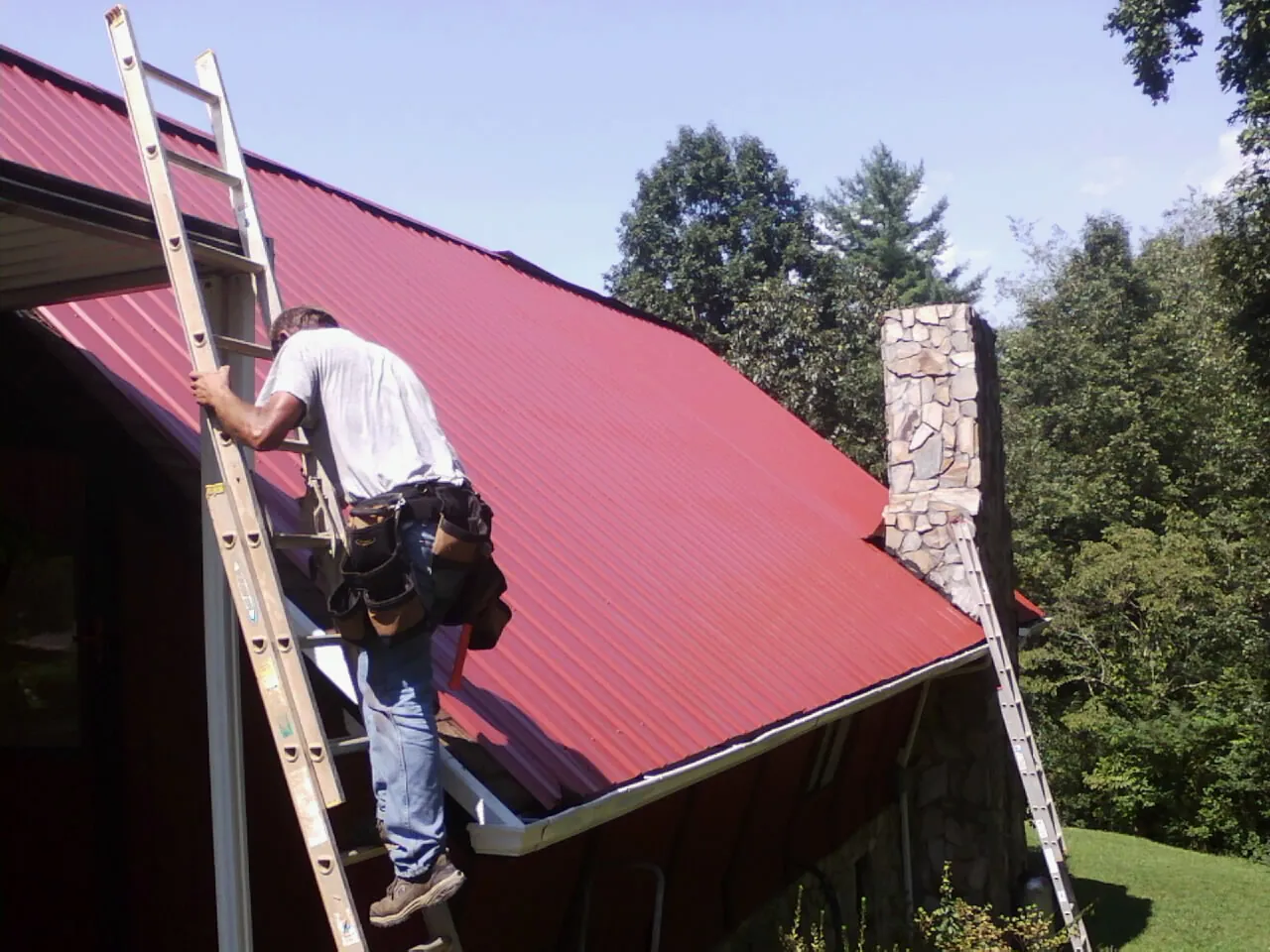 Roofer on a ladder beside a red metal roof and stone chimney — Soffit & Fascia Repair by Roofing Brothers in Oak Forest